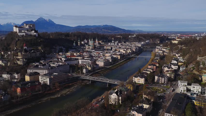 Aerial camera glides over Salzburg, Austria, revealing Hohensalzburg Fortress, Salzburg Cathedral, the Salzach River bridges, and the Alps in soft low daylight.