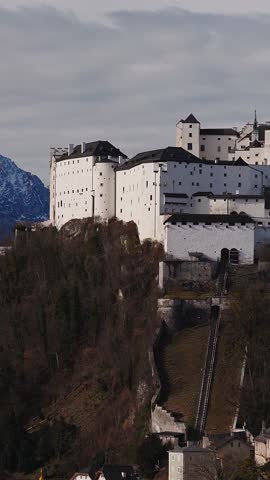 Aerial drone shot reveals Hohensalzburg Fortress, funicular tracks, and a red onion domed church above Salzburg, Austria, in cool daylight under banded clouds.
