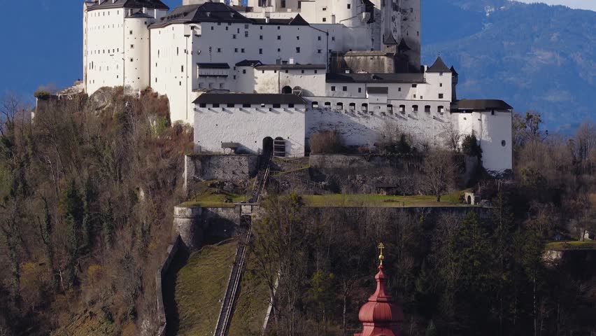 Aerial drone moves slowly toward Hohensalzburg Fortress in Salzburg, Austria, showing white walls, towers, battlements, funicular track, and snow capped Alps