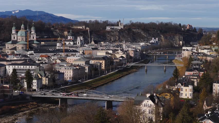 Aerial view of Salzburg, Austria shows the Salzach River, Salzburg Cathedral on the left, Hohensalzburg on a hill, arched bridges, a red tram, cars, and a gentle downstream flow.
