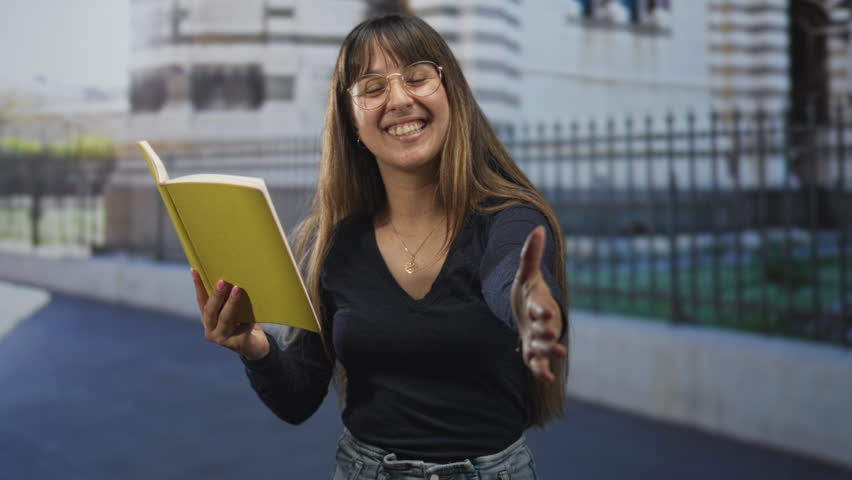 Woman holding yellow book and extending hand while smiling in front of building, reading to camera; friendly invitation joy.