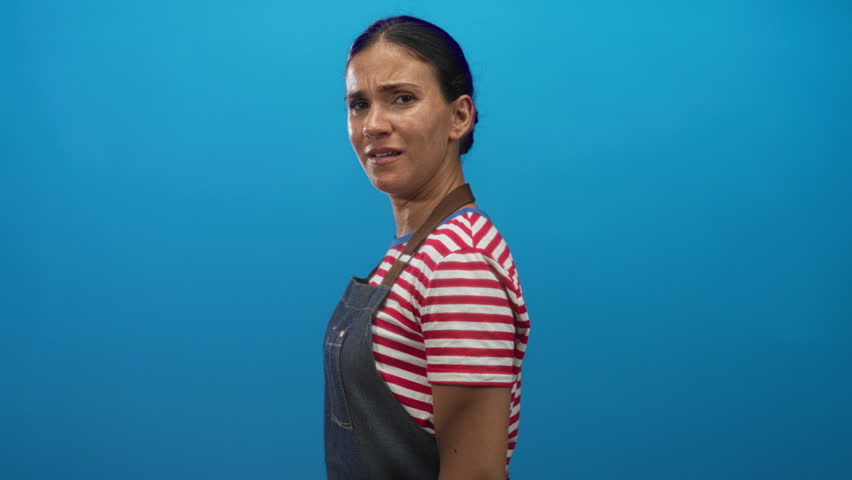 Woman in denim apron and red striped shirt rubbing her eyes and wiping tears in blue studio; sadness and vulnerability.
