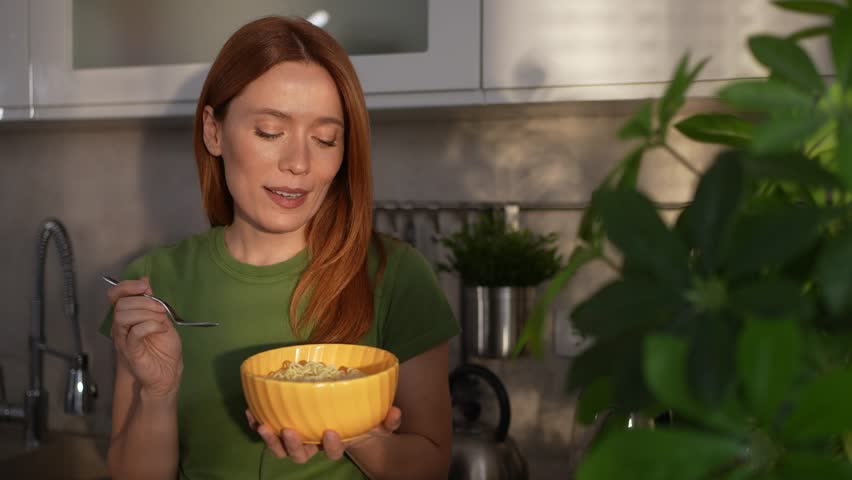 Beautiful smiling woman with red hair standing in the kitchen and eating delicious instant noodles from a yellow bowl for lunch, enjoying her meal with a happy and satisfied expression