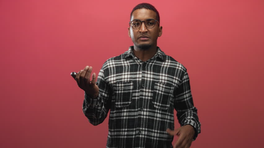 Man holding car keys, hands to mouth gesture, wearing plaid shirt against red studio backdrop; anxiety doubt uncertainty.