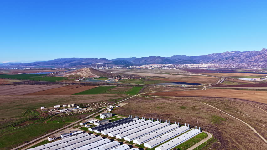Aerial view shows open fields and buildings in Montana region with mountains in distance under clear blue sky. Land is mostly flat with some agricultural plots and infrastructure visible.