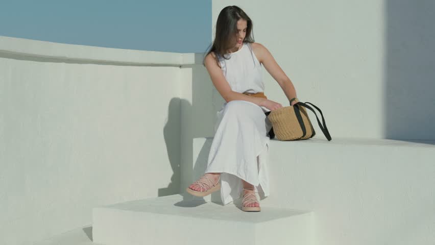 A young woman wearing a flowing white dress and stylish sandals sits outdoors with a woven straw bag, enjoying a bright, sunny day