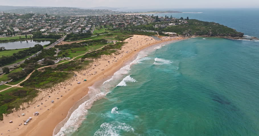 Aerial view of long sandy Curl Curl beach in Sydney with swimmers and sunbathers, turquoise ocean waves breaking on shore, green parkland, and residential neighborhoods of the northern beaches suburbs