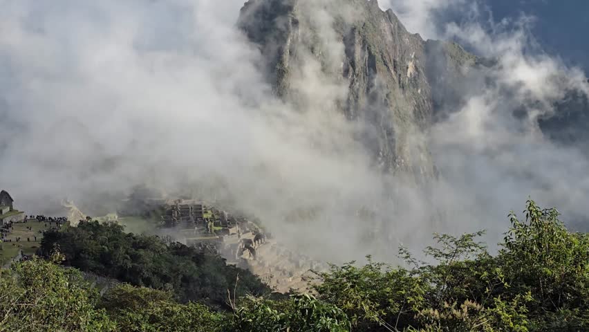 Machu Picchu, Peru - 26 June 2024. Clouds cover the ancient Inca site, obscuring terraces and stone buildings below a towering mountain, with dense greenery in front.
