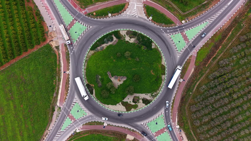 Top down aerial view of cars and trucks moving through a large roundabout with landscaped center island, lane markings and surrounding rural roads
