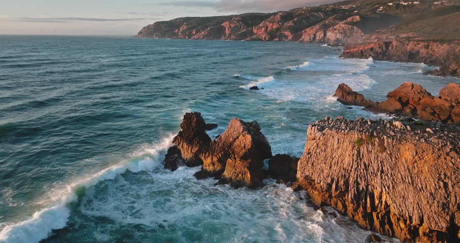 Atlantic Ocean waves crashing against the rugged cliffs and rock formations of Portugal's Sintra Cascais Natural Park coastline during a sunset with warm light illuminating the landscape. Drone flight