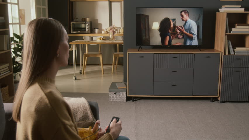 Medium full side shot of young Caucasian woman sitting down on couch with bowl of chips, watching love drama on flat screen digital TV, couple violently arguing and fighting