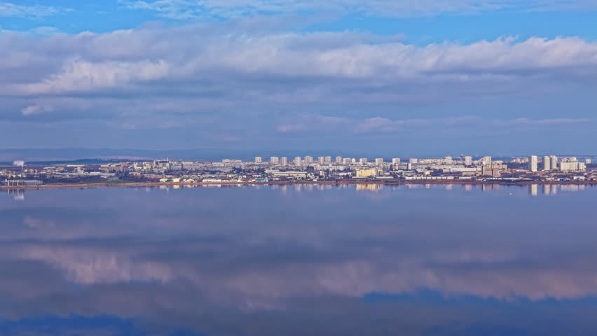 A city skyline shows modern buildings and structures along the waterfront. The clouds reflect in the water below, creating a unified view. The scene is bright and clear, revealing the landscape.