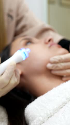 Professional cosmetologist in a spa salon performing a rejuvenating facial procedure with a modern hydrafacial device, using blue light therapy for skin cleansing and anti aging care