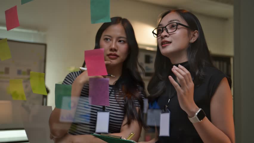 Asian businesswomen discussing strategy using sticky notes on a glass wall in office