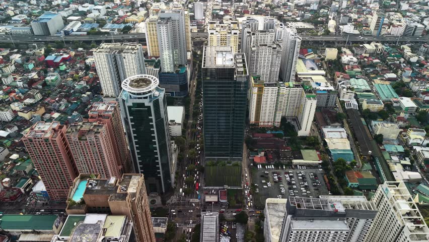 The camera drifts at high altitude in an oblique top-down angle over the Makati central business district in Manila, Philippines, revealing a wide multi-lane boulevard that cuts through two rows of towering high-rise buildings. In the foreground a distinctive tower with a circular rooftop helipad or observation deck anchors the left side of the frame, its curved architecture contrasting with the angular glass facades of neighboring corporate structures. Below, open parking lots filled with vehicles and a green sports court occupy the gaps between buildings, while the boulevard itself carries steady streams of traffic in both directions. The midground extends into a dense tapestry of residential quarters with low-rise rooftops in muted tones of gray, beige, and rust stretching outward from the commercial core. Overcast skies produce soft, even illumination that eliminates harsh shadows and allows uniform detail across the entire urban landscape, from reflective glass panels on the skyscrapers to individual cars on the roads below. The color palette leans toward cool grays and greens with occasional warm accents from terracotta rooftops and building facades. An elevated highway overpass is visible in the far background, tracing a curved path above the residential sprawl and reinforcing the layered infrastructure of this metropolitan area. The overall mood conveys the organized density and vertical ambition of a Southeast Asian financial hub surrounded by the organic spread of everyday city life.