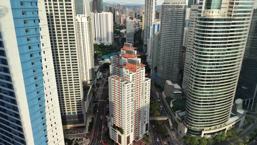 The camera moves forward at rooftop level, gliding between tightly clustered skyscrapers that form a narrow urban canyon in the Makati central business district. A prominent white-and-red facade residential tower occupies the center of the frame, flanked by glass-clad office buildings and modern condominiums rising on both sides. Below, a multi-lane street cuts through the base of the buildings, with vehicles and green patches of landscaped courtyards visible between the structures. The midground reveals additional rows of towers extending toward the southern part of Metro Manila, while the background dissolves into a hazy horizon where mountains are faintly outlined against an overcast sky. Soft diffused daylight wraps around the facades, eliminating harsh shadows and producing an even tonal range across concrete, glass, and painted surfaces. The color palette leans toward muted warm grays and creams, accented by the terracotta-red panels on the central building and scattered green canopy below. A cylindrical glass tower on the right side of the frame adds a curved geometric counterpoint to the predominantly rectangular silhouettes. The overall mood conveys the dense vertical urbanization and economic energy of one of Southeast Asia