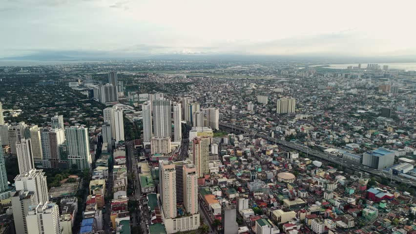 The drone captures a wide high-altitude panorama above the Makati Central Business District, tilting at an oblique downward angle as it slowly drifts laterally across the urban landscape. Tall residential and commercial towers dominate the left portion of the frame, their glass and concrete facades rising above a patchwork of lower structures that stretch endlessly toward the horizon. An elevated expressway carves a diagonal line through the mid-frame, separating the dense high-rise cluster from the sprawling low-rise neighborhoods that fill the center and right side of the composition. In the far background, the hazy outline of Manila Bay meets an overcast sky, adding depth and a sense of immense scale to the metropolitan area. The diffused cloud cover creates even, soft lighting with minimal harsh shadows, rendering the cityscape in muted warm tones of beige, gray, and faded green from scattered tree canopies. Rooftops of varying colors and materials — corrugated metal, concrete slabs, and painted surfaces — create a textured mosaic across the residential zones. The contrast between the orderly vertical towers of Makati and the organic horizontal density of surrounding neighborhoods illustrates the layered urban fabric of Metro Manila. Traffic flows along the expressway and narrow streets below, adding subtle movement to the otherwise static urban tableau.