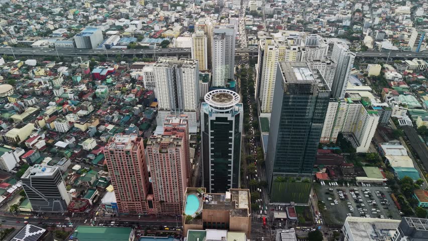 The drone hovers at an oblique downward angle above the transitional edge of the Makati Central Business District, where modern high-rise towers give way to dense low-rise residential neighborhoods. On the left side of the frame, a tall residential tower in warm terracotta tones features a rooftop swimming pool that glows turquoise against the surrounding concrete surfaces. The center of the composition is dominated by a dark-clad building topped with a distinctive circular architectural element, creating an immediately recognizable landmark within the urban fabric. To the right, a cluster of modern glass and concrete towers rises along a wide tree-lined boulevard that runs vertically through the frame, serving as a clear boundary between the commercial district and the residential areas beyond. In the background, an elevated highway stretches horizontally across the upper portion of the frame, with dense low-rise housing spreading outward beneath it in a seemingly endless patchwork of corrugated rooftops, narrow streets, and small structures. Overcast skies produce flat, even illumination that eliminates harsh shadows and allows architectural details to remain visible across all planes of the image. The color palette is restrained, dominated by grays, muted greens from street-level vegetation, and the occasional warm accent from painted facades. Parked vehicles dot the streets and parking areas near the base of the towers, indicating the scale of daily activity in this transitional urban corridor. The contrast between vertical modern development and horizontal informal density captures the characteristic layered growth pattern of Metro Manila.