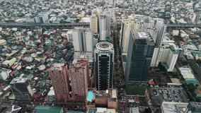 The drone hovers at an oblique downward angle above the transitional edge of the Makati Central Business District, where modern high-rise towers give way to dense low-rise residential neighborhoods. On the left side of the frame, a tall residential tower in warm terracotta tones features a rooftop swimming pool that glows turquoise against the surrounding concrete surfaces. The center of the composition is dominated by a dark-clad building topped with a distinctive circular architectural element, creating an immediately recognizable landmark within the urban fabric. To the right, a cluster of modern glass and concrete towers rises along a wide tree-lined boulevard that runs vertically through the frame, serving as a clear boundary between the commercial district and the residential areas beyond. In the background, an elevated highway stretches horizontally across the upper portion of the frame, with dense low-rise housing spreading outward beneath it in a seemingly endless patchwork of corrugated rooftops, narrow streets, and small structures. Overcast skies produce flat, even illumination that eliminates harsh shadows and allows architectural details to remain visible across all planes of the image. The color palette is restrained, dominated by grays, muted greens from street-level vegetation, and the occasional warm accent from painted facades. Parked vehicles dot the streets and parking areas near the base of the towers, indicating the scale of daily activity in this transitional urban corridor. The contrast between vertical modern development and horizontal informal density captures the characteristic layered growth pattern of Metro Manila. - Powered by Shutterstock - Get 15% off with code: PIKWIZARD15