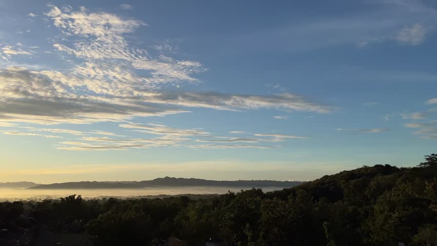 Sunrise sky with soft golden light illuminating clouds above a distant forest horizon, filmed during early morning in a peaceful natural landscape. The footage shows who: natural environment without people, what: sun rising behind clouds over tree line, when: early morning at sunrise, where: rural or forest landscape, and why: representing hope, new beginnings, calm atmosphere, and natural beauty. The scene conveys a warm, inspiring, and tranquil mood ideal for emotional storytelling.