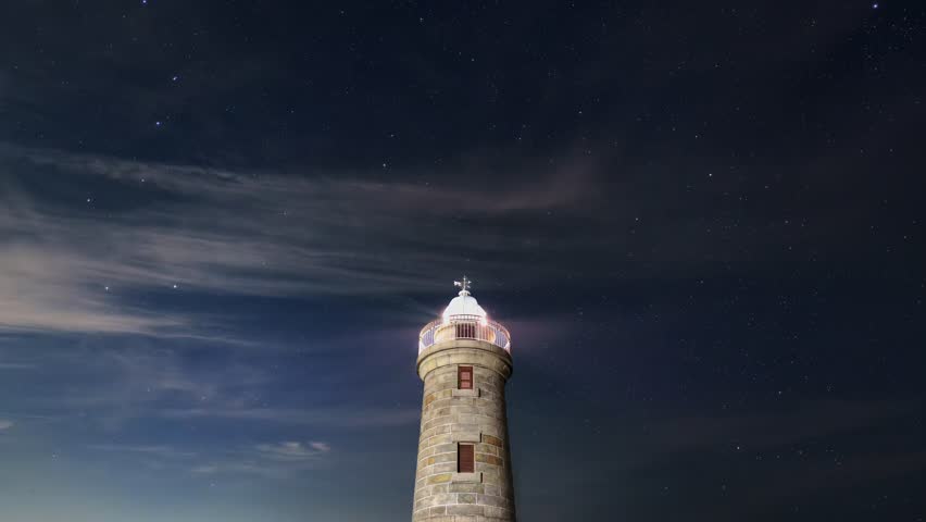 Historic stone lighthouse at night with a glowing lantern under a starry sky and wispy clouds.