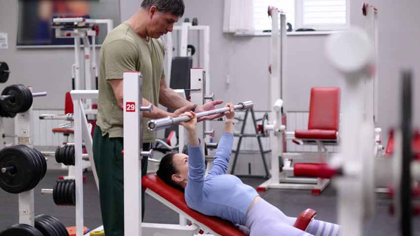 Slow motion video of a bench press under the supervision of a trainer. A Caucasian woman trains her chest muscles in a gym under the supervision of a trainer. Gym workout 4k footage.