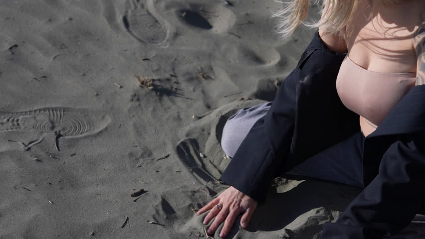 Close-up of a blonde woman sitting on the sand, picking it up, and letting it run through her fingers. Concept of the passage of time