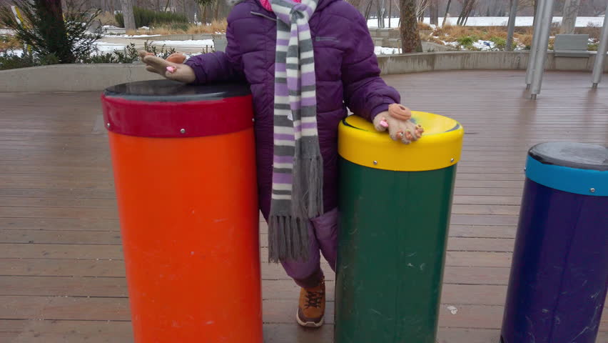 A young girl in a purple winter jacket and scarf plays on vibrant outdoor percussion instruments at the House of Music Hungary, exploring rhythm in a public park setting.