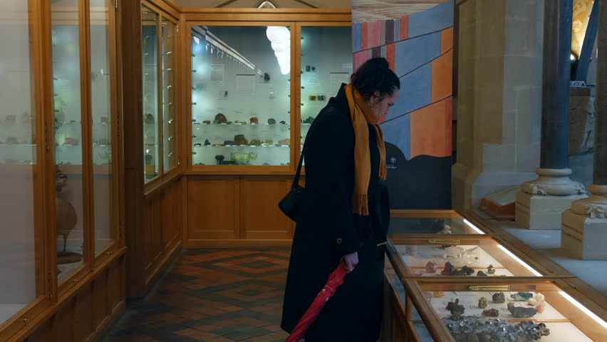 A woman examining mineral specimens inside a museum display case, holding a closed red umbrella while standing beside illuminated exhibits in a historic natural history gallery