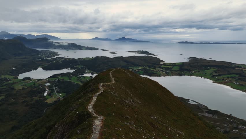 Hiking path with stone slabs following ridge line above fjords and islands under cloudy sky, Norway