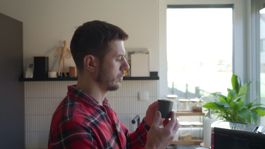 Man in red pajamas holding smelling coffee by window in kitchen interior. Person drinking cup of fresh espresso in the morning