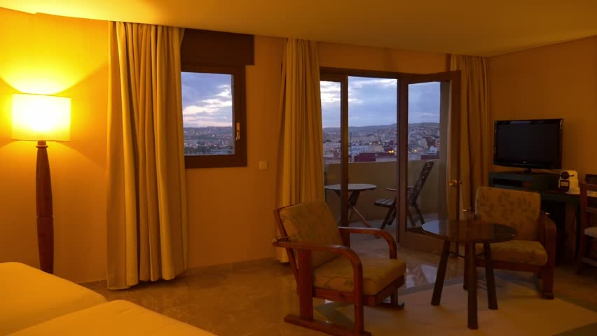 Tracking shot from a warm hotel room interior through open balcony doors to a panoramic city view of Melilla. Twilight sky, urban lights, and cozy furniture. Hospitality and travel concept.
