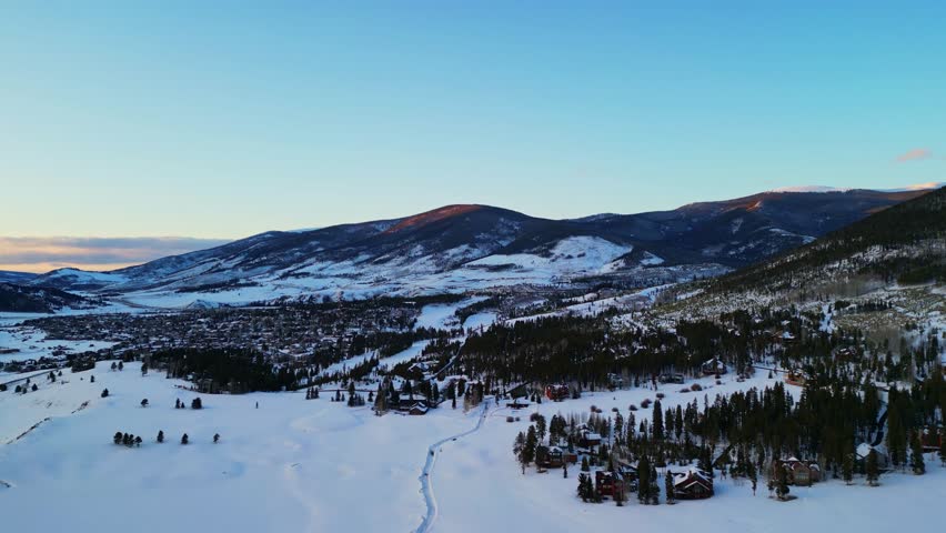 A wide aerial shot of a snow-covered mountain landscape featuring a small town nestled among pine forests as the sun hits the peaks at sunrise.