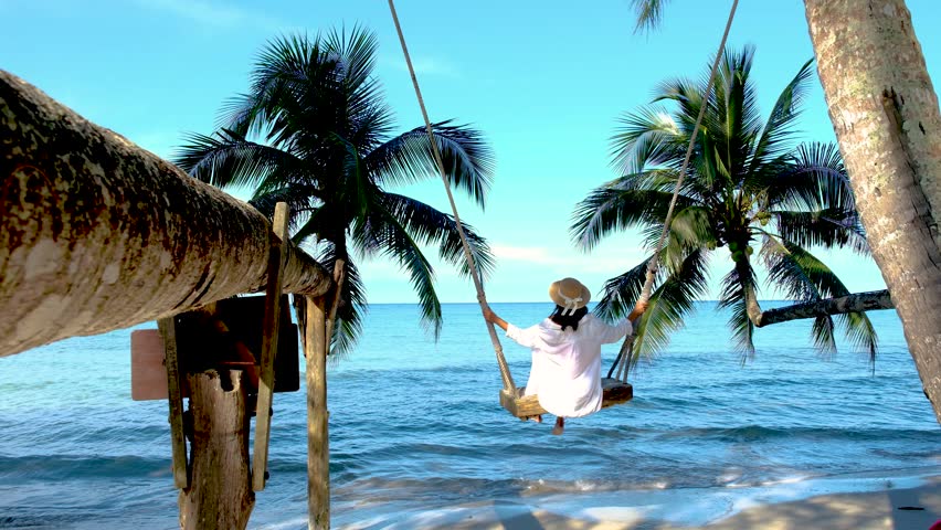 A serene moment on Koh Kood Island, where a woman enjoys swinging gently by the beach surrounded by lush palm trees. The turquoise waters and clear skies create a picture-perfect tropical paradise.