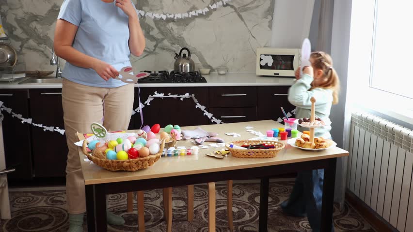 Wide shot of Caucasian mother and little daughter prepare for Easter by trying on a paper rabbit mask and playing together. Concept of Spring Festival