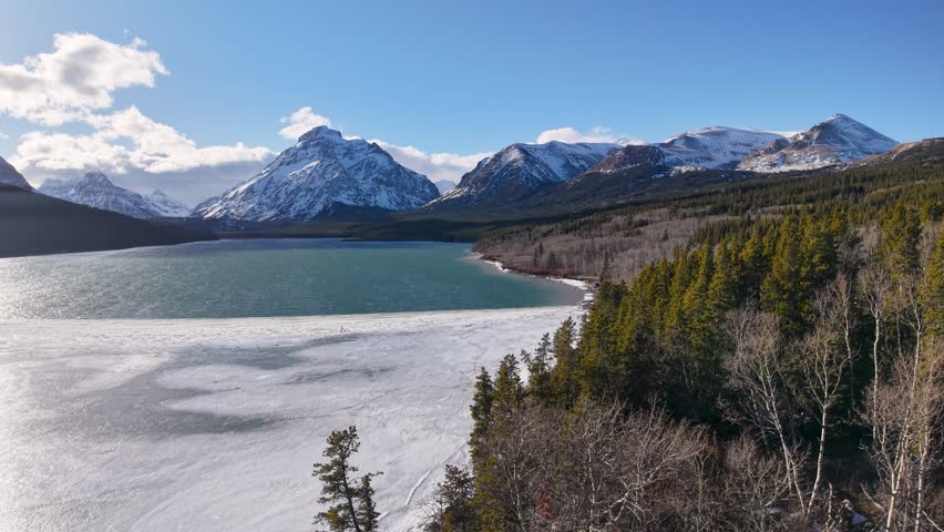 Aerial shot of a turquoise mountain lake partially covered by ice, bordered by dense conifer forest and snow-capped Rocky Mountains beneath a clear blue sky near Whitefish, Montana.