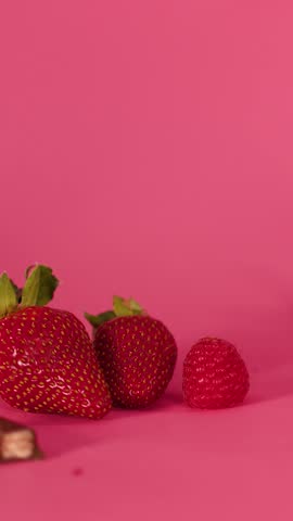 A hand arranges a sandwich cookie among strawberries, raspberries, and chocolate on a pink background.