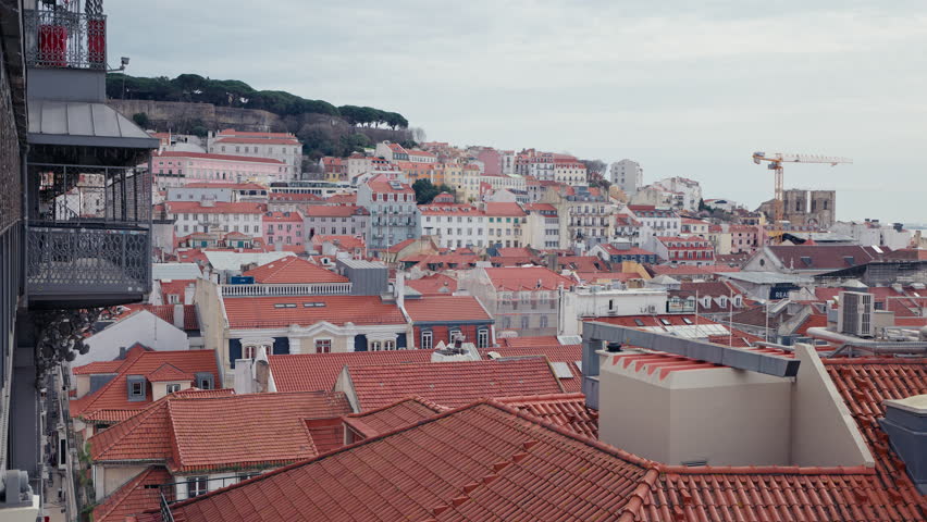A sweeping rooftop panorama of Lisbon, Portugal, framed by the iron structure of the Santa Justa elevator, with colorful hillside buildings and the Castelo de Sao Jorge visible under an overcast winte
