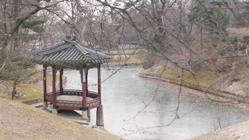 Slow motion landscape showing wooden temple hut resting near frozen lake with tree branch in foreground at Gwallamjeong pavilion inside Secret Garden Seoul city South Korea travel tourism and nature