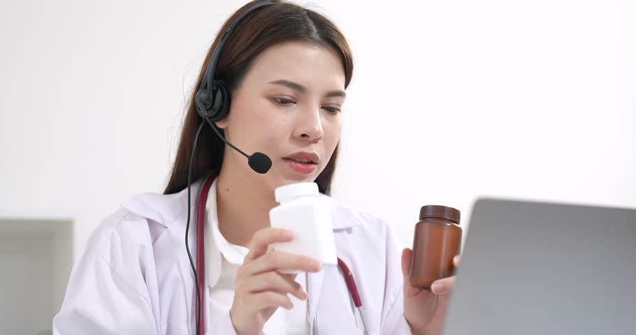 Asian female doctor consulting online using laptop and headset, demonstrating medicine bottles during telemedicine session, explaining dosage and health advice, modern healthcare communication