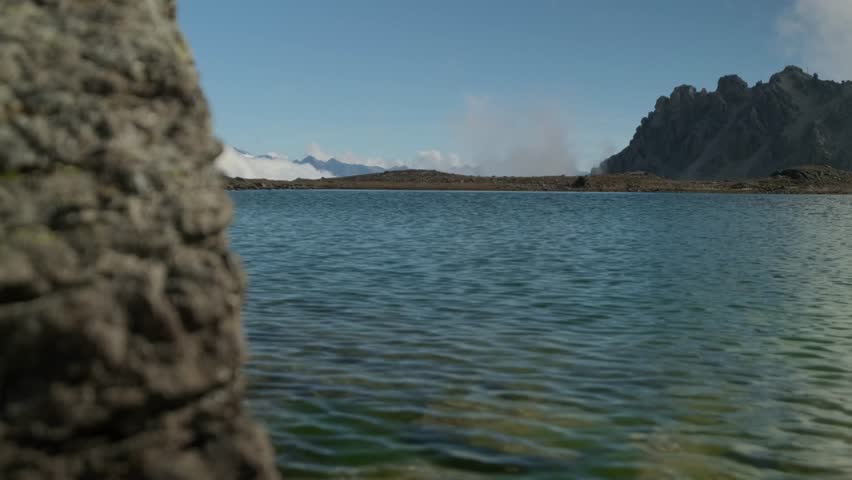 A tranquil alpine lake reflects the clear blue sky, with rugged mountains and wisps of clouds in the background, creating a peaceful natural landscape
