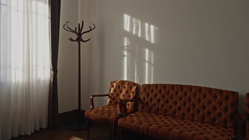 A serene interior view of a historic Western-style room featuring a tufted brown velvet sofa, a vintage coat rack, and soft sunlight filtering through lace curtains.