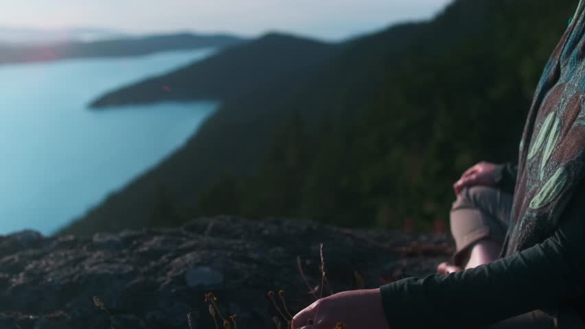 A lone individual finds peace and contemplation on a rocky precipice, gazing at the tranquil waters and distant peaks as daylight fades