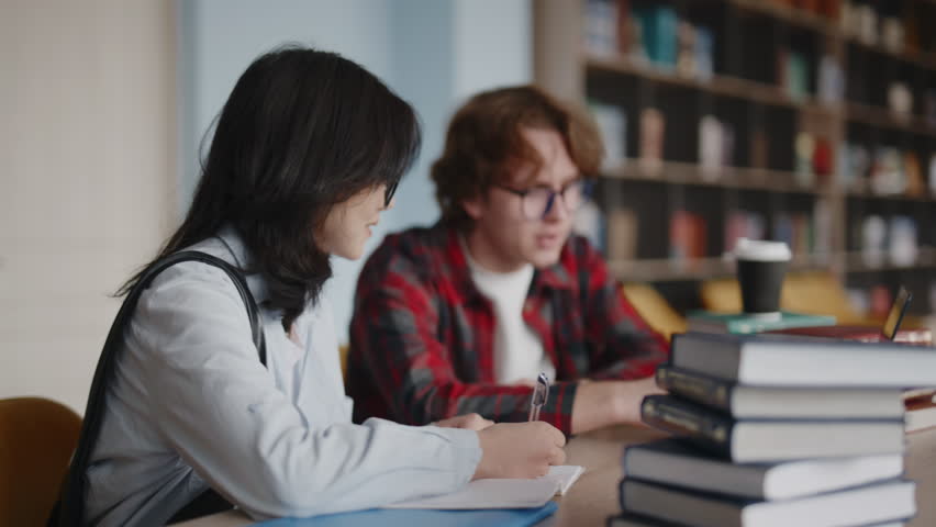 Diverse college students studying together in a campus library, working on a laptop, taking notes, and discussing their assignment with a stack of books on the desk for research and learning