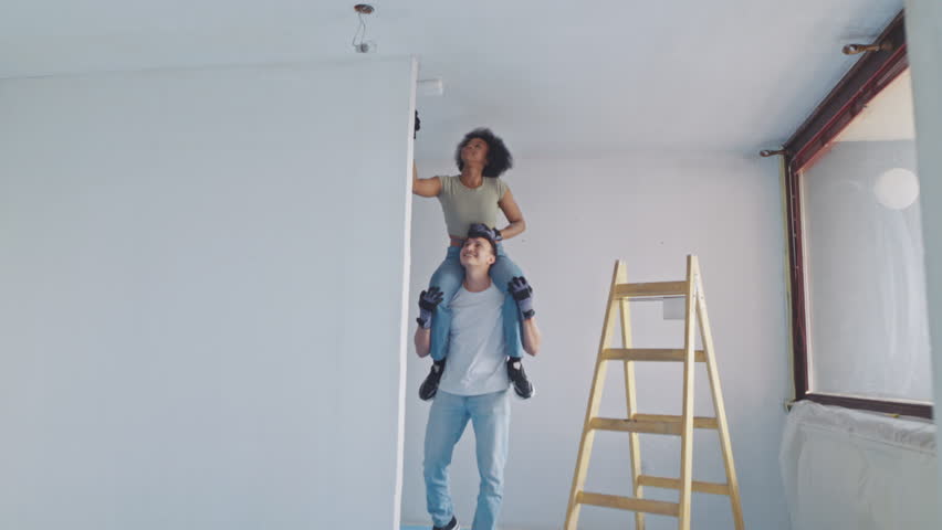 A cheerful couple collaborates on painting their home interior amidst a vibrant apartment renovation. The man supports the woman perched on his shoulders, both in casual attire.