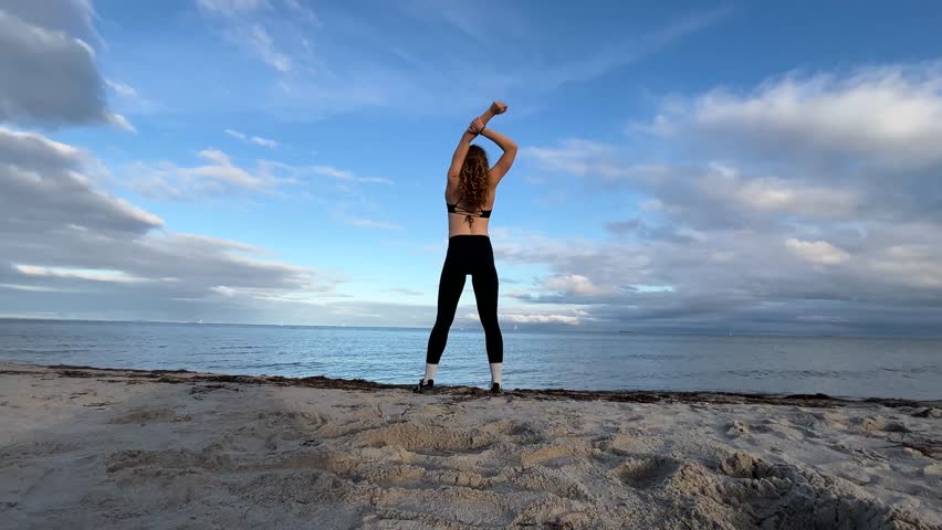 Fit young woman on beach stretching arms towards a blue sky – enjoying life, ocean, and nature