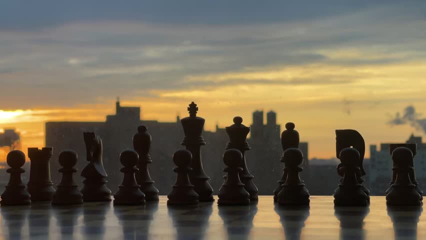 A pensive and cinematic silhouette of chess pieces against a vibrant sunset sky, reflecting strategic planning and competition. The high-contrast image creates a thoughtful mood and suggests a battle of minds.
