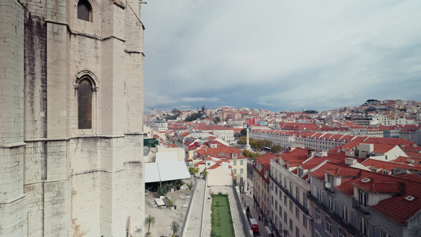 A sweeping elevated view from the Convento do Carmo in Lisbon, Portugal, with the Gothic church tower in the foreground and a vast panorama of terracotta rooftops stretching toward Rossio square under