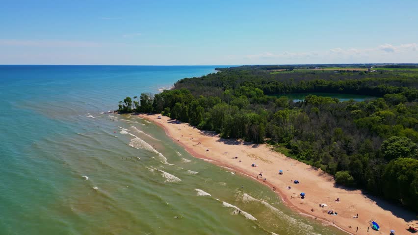 A stunning high-angle drone shot capturing the meeting of turquoise water, a sandy beach, and a dense green forest under a clear blue sky. Shot in Wisconsin.