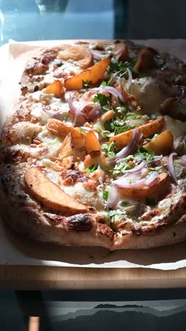 A freshly prepared pizza sits on a wooden surface, showcasing various toppings like fruit and vegetables. Natural light highlights the cheese and ingredients, suitable for culinary content.